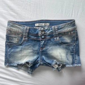 Lågmidjade jeansshorts - Snygga blå jeansshorts från Oversion med slitna detaljer, fransig kant och coola nitar i brons och turkos på fickorna. Shortsen har låg midja, tre knappar framtill och klassisk femficksdesign. Perfekta för sommaren och ger en edgy vibe. Ingen lapp men skulle säga S💕