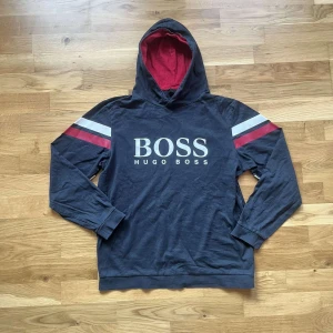 Hugo boss hoodie  - Svart hoodie från Hugo Boss med stort vitt BOSS-tryck på bröstet. Röd och vit rand på ärmarna ger en sportig vibe. Mjuk insida och huva med röd fodring. Långärmad och i bomull, perfekt för chill dagar. Storlek L men sitter som S så kolla måtten noggrant 