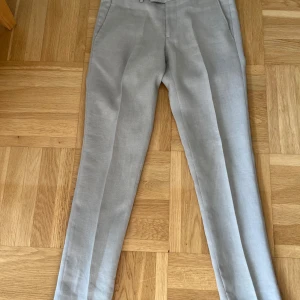 Beige linnekostymbyxor J.Lindeberg - Snygga beige kostymbyxor från J.Lindeberg i linne. Byxorna har raka ben, pressveck, bälteshällor och diskreta bakfickor. Perfekta för en clean och stilren look under varmare dagar. Använd 1 ggr. Nyskick! 