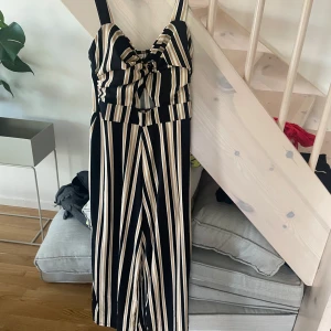 Randig långklänning från Amisu - Snygg långklänning från Amisu med svarta, beige och vita ränder. Klänningen har smala axelband, knytdetalj framtill och en figurnära överdel. Perfekt för dig som gillar en trendig och stilren look.