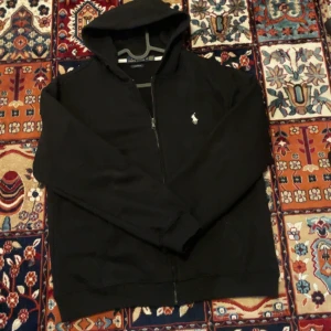 Svart hoodie från Polo Ralph Lauren - Svart hoodie från Polo Ralph Lauren i custom fit med dragkedja framtill och klassisk broderad logga på bröstet. Tröjan har huva och långa ärmar, perfekt för en chill och stilren look. Materialet är mjukt och skönt.