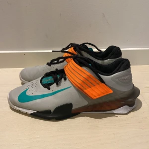 Tyngdlyftarskor Nike Savaleos träningsskor grå/orange strl 42,5 - Nike Savaleos träningsskor i grått med turkos swoosh och detaljer, samt en bred orange rem över vristen. Skorna har svart meshfoder, snörning och platt sula för stabilitet. Perfekta för gymmet och tyngdlyftning. Syntetmaterial och sportig look.