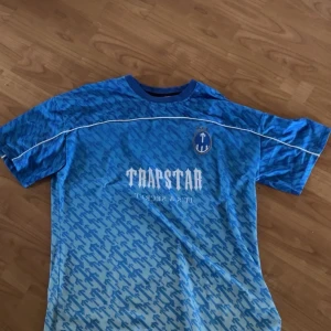 Blå Trapstar fotbollströja - Snygg blå fotbollströja från Trapstar med vit logga och nummer 22 på ryggen. Tröjan har ett unikt mönster över hela tyget och vita detaljer längs axlarna. Tillverkad i lätt och ventilerande material, perfekt för dig som gillar streetwear och sportig stil. Lite större i storleken rekommenderar folk med L eller dom som gillar lite baggy