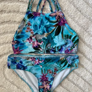 Blommig bikini från KappAhl i blått - Endast använd en gång, så nyskick! Superfin bikini från KappAhl med tropiskt blommönster i blå, lila och gröna toner. Hög halsringning och smala axelband som korsas i ryggen. Materialet är mjukt och stretchigt, perfekt för strandhäng. Färgglada blommor och papegojor ger en riktigt somrig vibe.
