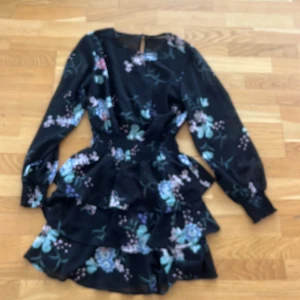 Blommig volangklänning från Gina Tricot - Supersöt svart kortklänning med blommigt mönster i blått, lila och rosa från Gina Tricot. Klänningen har långa ärmar med smock vid handlederna, markerad midja och flera volanger i kjolen. Perfekt för dig som gillar romantisk och trendig stil.