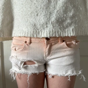 Ljusrosa och vita jeansshorts med slitningar - Midjemått 37.                                                     Snygga jeansshorts i en ljusrosa till vit ombre med råa, fransiga kanter och slitna detaljer framtill. Klassisk femficksmodell med knapp och dragkedja. Perfekta för en chill sommarlook.