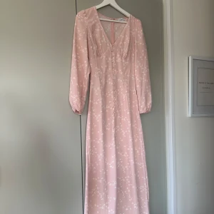 Rosa klänning från Nelly - Säljer en  supersöt rosa långklänning från Nelly X GLAMOROUS med vita blommor på. Klänningen har långa ärmar, v-ringning, knappar framtill och slit nertill. Svalt material så perfekt för vår och sommar! Endast använd 2 gånger, säljer pga för liten!