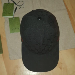 Svart keps från Gucci - Snygg svart keps från Gucci med det klassiska GG-mönstret. Kepsen har en justerbar rem baktill och detaljer i rött och grönt på sidan. Perfekt för en stilren look.