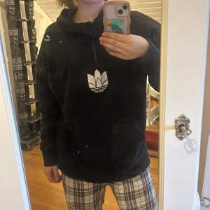 Svart hoodie från Adidas - Säljer en svart hoodie från Adidas med det klassiska trefoil-loggan i vitt på bröstet. Hoodien har en dragkedja vid halsen och en stor ficka framtill.