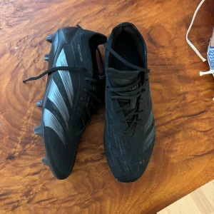 Svarta fotbollsskor  Adidas predator - (Nypris 3000kr) Säljer ett par svarta fotbollsskor från Adidas med snörning och mönstrade detaljer i silver. Skorna har dobbar för bra grepp på planen och en modern design som passar perfekt för fotbollsträning och matcher.