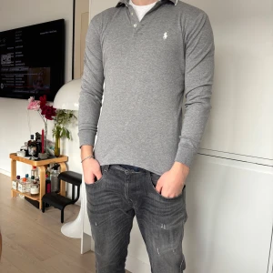 Grå pikétröja från Ralph Lauren - Snygg grå pikétröja från Ralph Lauren med klassisk krage och knappar. Tröjan har långa ärmar och en liten broderad logga på bröstet. Perfekt för en stilren look.