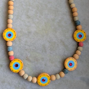 Färgglatt halsband med träpärlor - Unikt halsband med träpärlor i olika färger som beige, blå, rosa och gult. Halsbandet har runda, målade träplattor med cirkulära mönster i gult, blått och rött. Perfekt för att ge en färgglad touch till din stil.