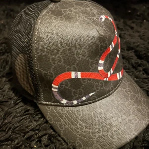 Svart keps  - Snygg svart keps från med ett coolt ormmönster i rött och vitt. Kepsen har ett justerbart band bak med röd och blå rand. Perfekt för att ge din outfit en lyxig touch.