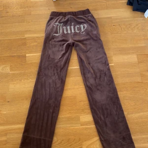 Bruna byxor från Juicy Couture - Snygga bruna byxor från Juicy Couture i mjukt material med glittrande text på baksidan. Perfekta för en avslappnad stil med en touch av glamour. Byxorna har en elastisk midja och dragsko för extra komfort. Endast använda ett fåtal gånger och är i mycket bra skick!💗