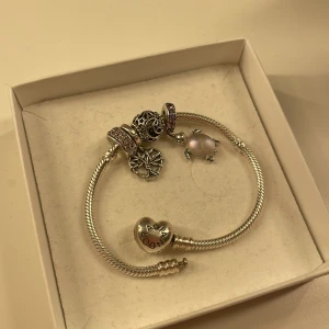 Silverarmband med berlocker från Pandora - Elegant silverarmband från Pandora med flera unika berlocker. Inkluderar en sköldpadda med rosa och lila detaljer, en berlock med rosa stenar, och en stjärn- och måndekorerad berlock. Perfekt för att ge en personlig touch till din stil. Sammanlagt värde med armband och berlocker är ca 3000kr.🤗 Skriv för fler bilder, ask och låda medföljer! Använt ca fem gånger!😊Pris går att diskutera vid snabb affär!🤗