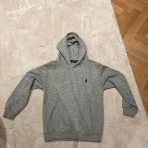 Grå hoodie från Ralph Lauren - Säljer en stilren grå hoodie från Ralph Lauren med det klassiska logotypen på bröstet. Tröjan har en bekväm passform och är perfekt för en avslappnad look. Den är tillverkad i mjukt material och har långa ärmar samt en praktisk huva. Pris kan diskuteras bytten med! Aldrig använd säljer på grund av att de är för små!