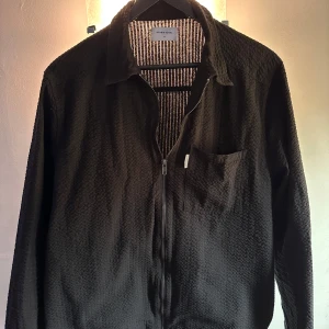 Svart jacka från studio total - Säljer en stilren svart jacka från Studio Total med dragkedja och en bröstficka. Jackan har en krage och är perfekt för en avslappnad look. Den har en diskret textur som ger den lite extra stil.