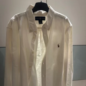 Vit skjorta från Ralph Lauren - Klassisk vit skjorta från Ralph Lauren med broderad logotyp på bröstet. Skjortan har långa ärmar och knappar framtill. 
