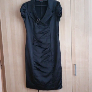 Svart klänning från Modal - Elegant svart klänning från Modal med korta ärmar och en v-ringad design. Klänningen har en dekorativ broschdetalj framtill och en dragkedja på sidan för enkel påklädning. Perfekt för en stilren look.