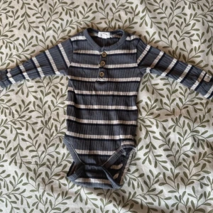 Randig body från Newbie - Säljer en söt randig body från Newbie i blå och grå nyanser. Bodyn har långa ärmar och tre knappar framtill för enkel påklädning. Perfekt för de små under kyligare dagar.