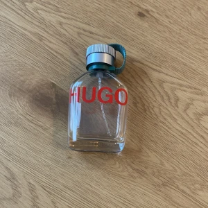 HUGO parfymflaska - Säljer en stilren parfymflaska från HUGO med en modern design. Flaskan är genomskinlig med röd text och har en grå kork med en grön rem. Perfekt för den som vill ha en fräsch och tidlös doft.