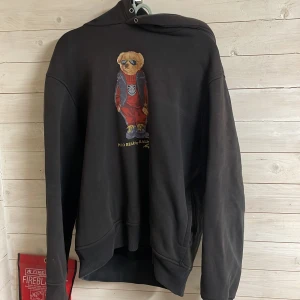 Svart hoodie från Polo Ralph Lauren - Säljer en svart hoodie från Polo Ralph Lauren med en ikonisk björn på framsidan. Tröjan har en klassisk passform med långa ärmar och en bekväm huva. Perfekt för en avslappnad stil. 🐻