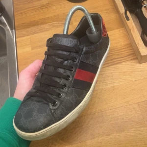 Gucci ace skor - Snygga svarta sneakers med diskret mönster och röd samt blå rand på sidan. Skorna har en röd detalj vid hälen och svarta snören. Perfekta för en stilren look.