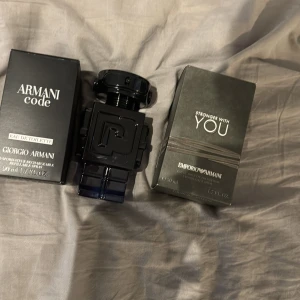 Parfymflaskor från Giorgio Armani - Två eleganta parfymflaskor från Giorgio Armani. Den ena är 'Armani Code' med en stilren svart förpackning och den andra är 'Stronger With You' i en mörkgrå förpackning. Flaskorna har en modern och lyxig design, perfekt för den som vill ha en sofistikerad doft.