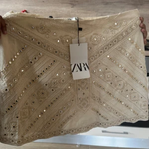 Beige kjol från Zara - Snygg beige kjol från Zara med glittriga paljetter och broderade detaljer. Kjolen är aldrig använd och prislappen är på. Den är köpt i Frankrike därav euro priset. Kjolen är i storlek S 