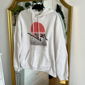 Vit hoodie med grafiskt tryck - Säljer en vit hoodie med ett coolt grafiskt tryck i rött och svart. Hoodien har en stor ficka framtill och justerbar huva med dragsko. Perfekt för en avslappnad stil.