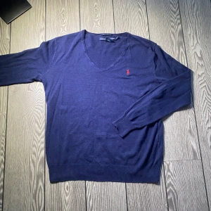 Ralph lauren - Säljer en stilren mörkblå tröja från Ralph Lauren med v-ringning och broderad röd logotyp på bröstet. Tröjan har långa ärmar och är tillverkad i ett mjukt material.