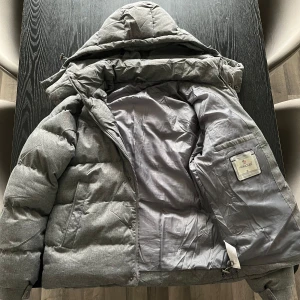 Grå dunjacka från Moncler - Säljer en snygg grå dunjacka från Moncler. Jackan har en quiltad design med en praktisk huva och dragkedja framtill. Perfekt för kyliga dagar. Den har även en broderad Moncler-logga på ärmen och tryckknappar på fickorna. Använd bara en gång. Hör av dig vid intresse!
