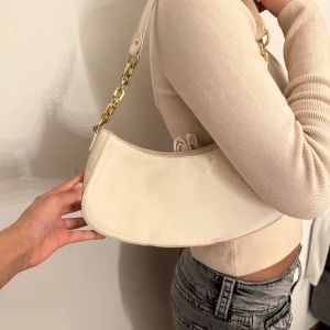 Beige axelväska med guldkedja - Snygg beige axelväska med en elegant guldkedja som axelrem. Väskan har en stilren och modern design med en glansig yta.