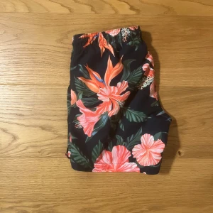 Blommiga badshorts från GANT - Snygga badshorts från GANT med ett färgglatt blommönster i rosa och grönt på en svart bakgrund. Shortsen har en elastisk midja med snörning för bästa passform. Perfekta för sommarens stranddagar!