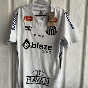 Vit fotbollströja från Umbro - Säljer en vit fotbollströja från Umbro med flera sponsormärken och nummer 10 på ryggen. Tröjan har korta ärmar och klubbmärke på bröstet. Perfekt för fotbollsfans!