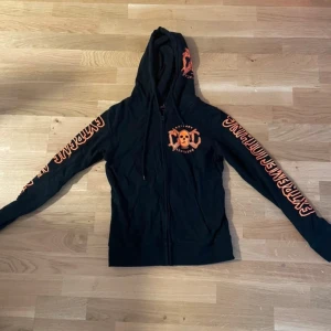 Svart hoodie från Extreme Clothing - Säljer en svart hoodie från Extreme Clothing med orange dödskalletryck och text på ärmarna. Hoodien har dragkedja och huva, perfekt för en cool och avslappnad stil. Passar bra för dig som gillar unika och edgy plagg.