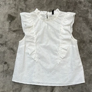 Vit blus med volangdetaljer - Säljer en söt vit blus med volangdetaljer och broderade hålmönster. Blusen är ärmlös och har en rund halsringning. Perfekt för en somrig look!