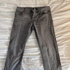 Grå jeans från Jack & Jones - Snygga grå jeans från Jack & Jones med en klassisk femficksdesign. De har en knappgylf och är perfekta för en stilren look. Passar bra till både vardag och mer uppklädda tillfällen. Passform Slim Glenn. Skriv för mer bilder eller frågor.