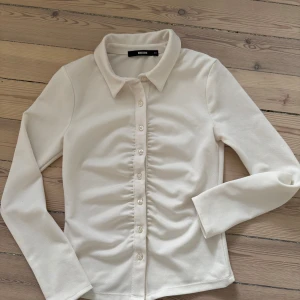 Vit långärmad skjorta från Bikbok - Säljer en stilren vit skjorta från Bikbok  med långa ärmar och knappar framtill. Skjortan har en klassisk krage och en lätt rynkad detalj längs knappraden. Perfekt för en chic och enkel look.