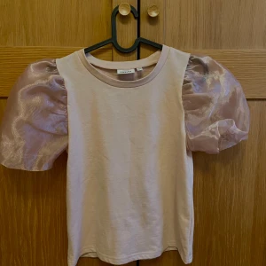 Rosa topp med puffärmar  - Säljer en rosa topp med unika puffärmar i ett glansigt material. Perfekt för att ge en extra touch till din outfit. Toppen har en rund halsringning och korta ärmar.