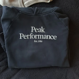 Blå/Svart hoodie från Peak Performance - Säljer en stilren svart hoodie från Peak Performance med vit logga på bröstet. Tröjan har en klassisk känguruficka och en bekväm huva. Perfekt för en avslappnad stil. Saknar snören.