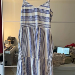 Vit och blå klänning med mönster - Säljer en somrig vit och blå klänning med smala axelband och ett vackert geometriskt mönster. Klänningen har en v-ringad hals och ett knytband i midjan för en fin passform. Perfekt för varma dagar!