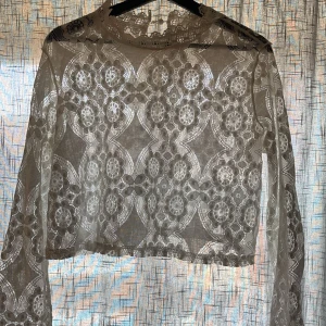 Vit spetsblus - Elegant vit spetsblus med blommigt mönster. Blusen har långa ärmar och en transparent design som ger en romantisk känsla. Perfekt för att lägga till en touch av elegans till din outfit.