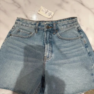 Blå högmidjade jeansshorts från Pull&Bear - Snygga blå jeansshorts från Pull&Bear med hög midja och klassisk femficksdesign. Shortsen har råa kanter nertill och bälteshällor. Perfekta för sommardagar och lätta att matcha med allt. Styla med en crop top eller hoodie för en chill look.