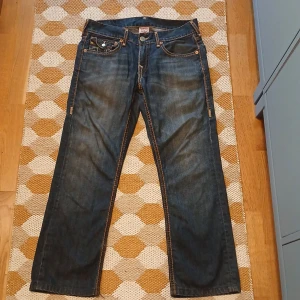True Religion jeans med kontrastsömmar - Säljer ett par blå jeans från True Religion med ikoniska kontrastsömmar och stora bakfickor med lock och knappar. Klassisk femficksmodell i denim med snygg tvätt och detaljerade sömmar. Perfekta för dig som gillar streetwear och vill sticka ut.