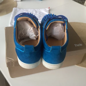 Blå Christian Louboutin sneakers med nitar - Snygga blå sneakers från Christian Louboutin i mocka med matchande blå snörning och coola nitar på tån. Skorna har den klassiska röda sulan och vita kanter. Insidan är i beige skinn för extra komfort. Perfekta för dig som vill sticka ut med din stil.