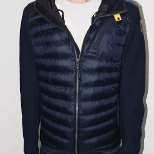Marin blå  pufferjacka från Parajumpers S - Säljer en maten blå pufferjacka från Parajumpers med huva och coolt badge på ärmen. Jackan har quiltad framsida, dragkedja och detaljer i mesh på ärmarna. Perfekt för kalla dagar och har en sportig vibe. Varan är i mycket bra skick och har inga tecken på användning