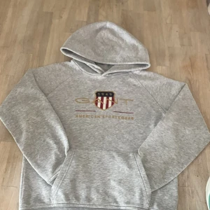 Grå hoodie från GANT med broderad logga - Snygg grå hoodie från GANT med broderad logga och sköld på bröstet. Klassisk känguruficka framtill och huva. Tillverkad i mjuk bomullsblandning, perfekt för chill dagar. Stilsäker och enkel att matcha med jeans eller joggers.
