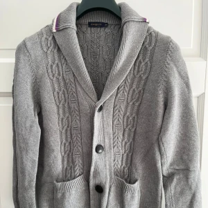 Grå stickad kofta från Henri Lloyd - Snygg grå stickad kofta från Henri Lloyd med kabelstickat mönster framtill, bred krage med lila och vit rand, samt stora knappar och två fickor. Perfekt för lager-på-lager och chill stil under kyliga dagar.