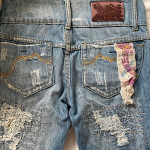 Lågmidjade bootcut/raka  jeans (trasig dragkedja) - Säljer ett par blå lågmidjade bootcut jeans med slitningar och lagningar, broderade detaljer på bakfickorna. De e från Miss Yang. Tyvärr är dragkedjan trasig (se bild 3+4), vilket kanske inte spelar så stor roll då de går att knäppa med de två knapparna som finns! I övrigt är de i jättefint skick💕står storlek 27, men jag som vanligtvis bär storlek s/w25 upplevde dem som lagom förutom att de är för tighta runt höfterna. Midja ett varv runt om : 72 cm Innerben: 82,5💕 Tar emot rimliga prisförslag